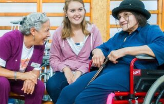 Mary Kay smiling at an elderly woman in a wheelchair and laughing and a young woman in the center of them smiling into the distance.
