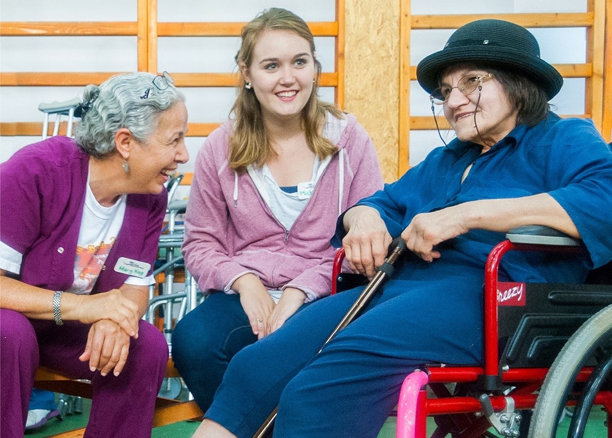 Mary Kay smiling at an elderly woman in a wheelchair and laughing and a young woman in the center of them smiling into the distance.