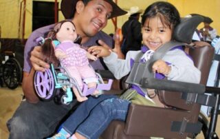Annadelce in her new wheelchair with a man next to her smiling at the camera as he holds a doll in a wheelchair.