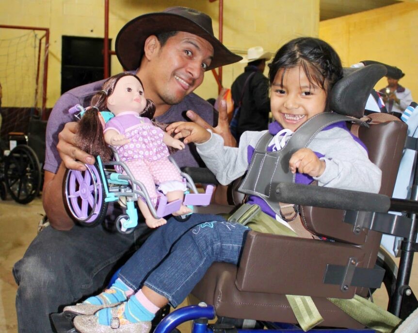 Annadelce in her new wheelchair with a man next to her smiling at the camera as he holds a doll in a wheelchair.