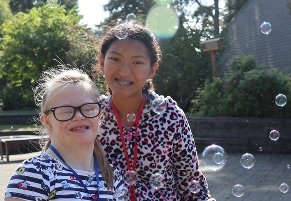 A little girl with Down Syndrome smiling with another little girl as they play with bubbles.