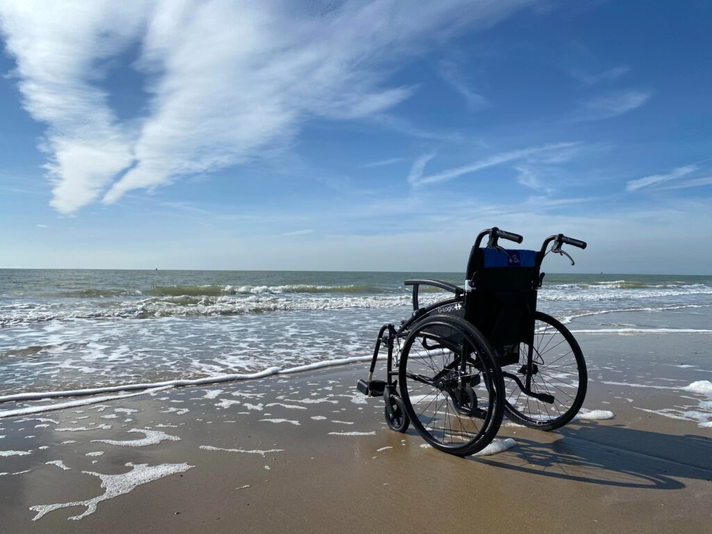 A wheelchair sitting on the beach facing the foamy waters of the ocean. A blue sky above.