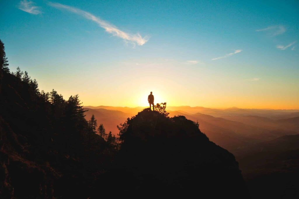 Silhouette of trees, rolling hills and man on top at sunrise.