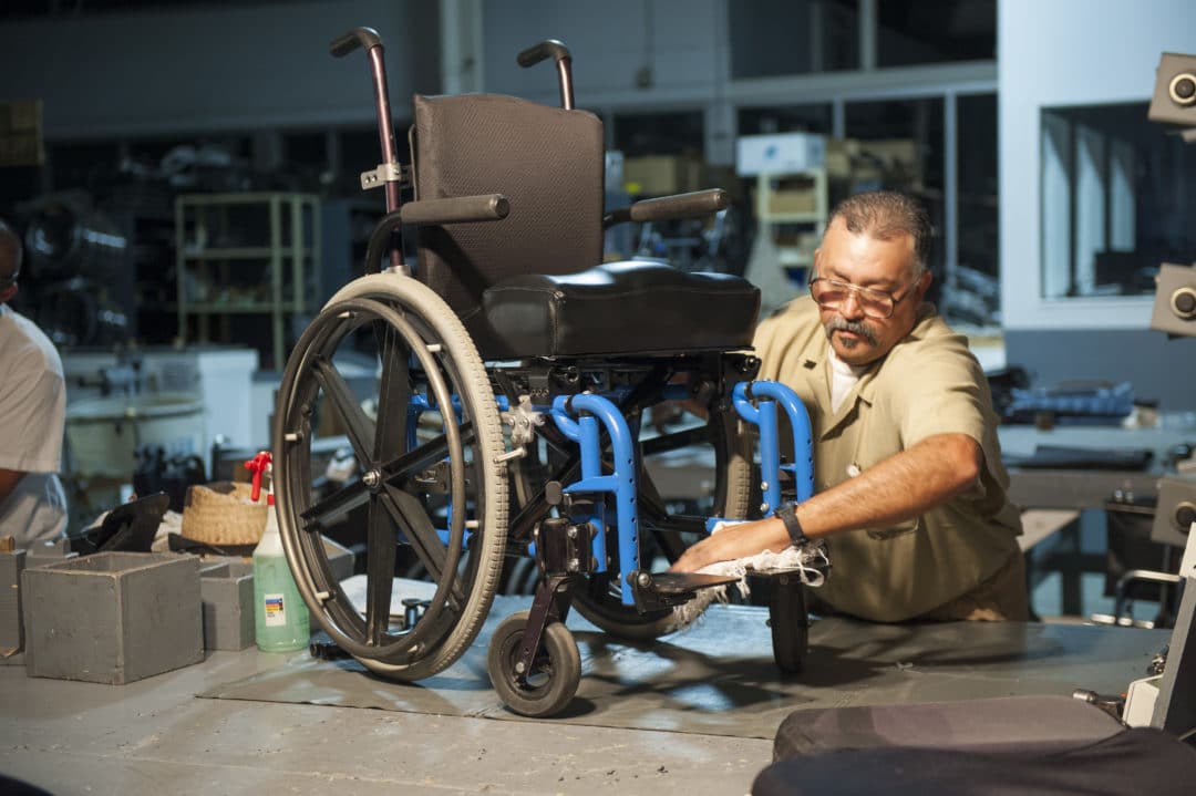A man cleaning a wheelchair and restoring it in a warehouse with all kinds of tools in it.