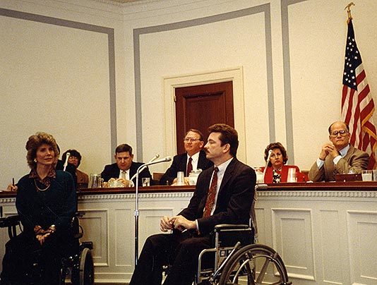 Joni Eareckson Tada sitting at the front of a council chamber speaking as she's seated in her wheelchair, sitting next to another man also using a wheelchair. Five other council members sit behind her behind a large circular desk with an American flag standing behind them.