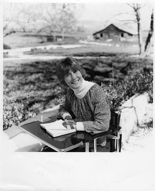 A historical black and white photo of Joni at her family farm in Maryland seated in her wheelchair with her bible in from of her as she's smiling at the camera.