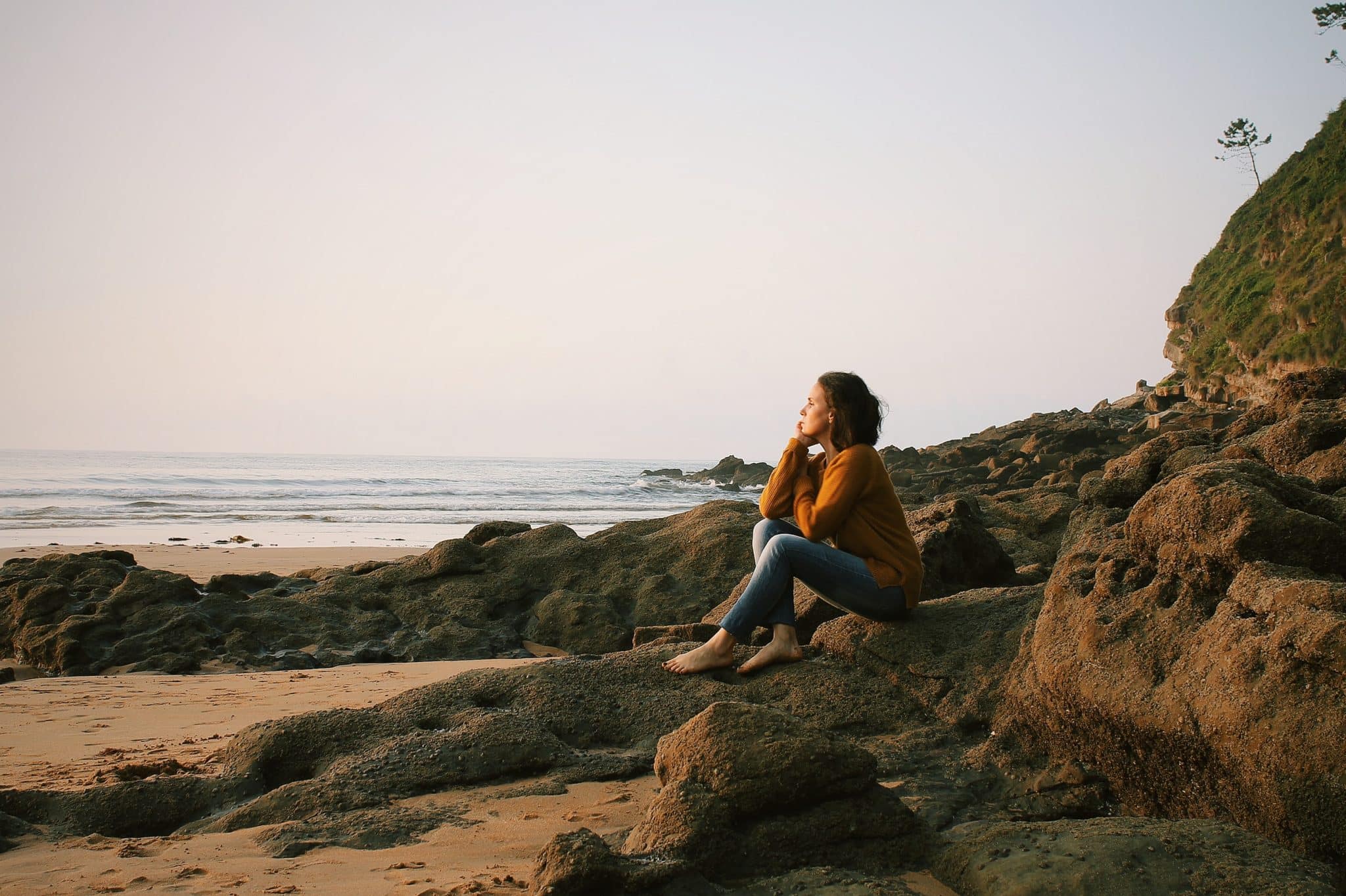 A woman sitting on a rock on the seashore looking out at the ocean.