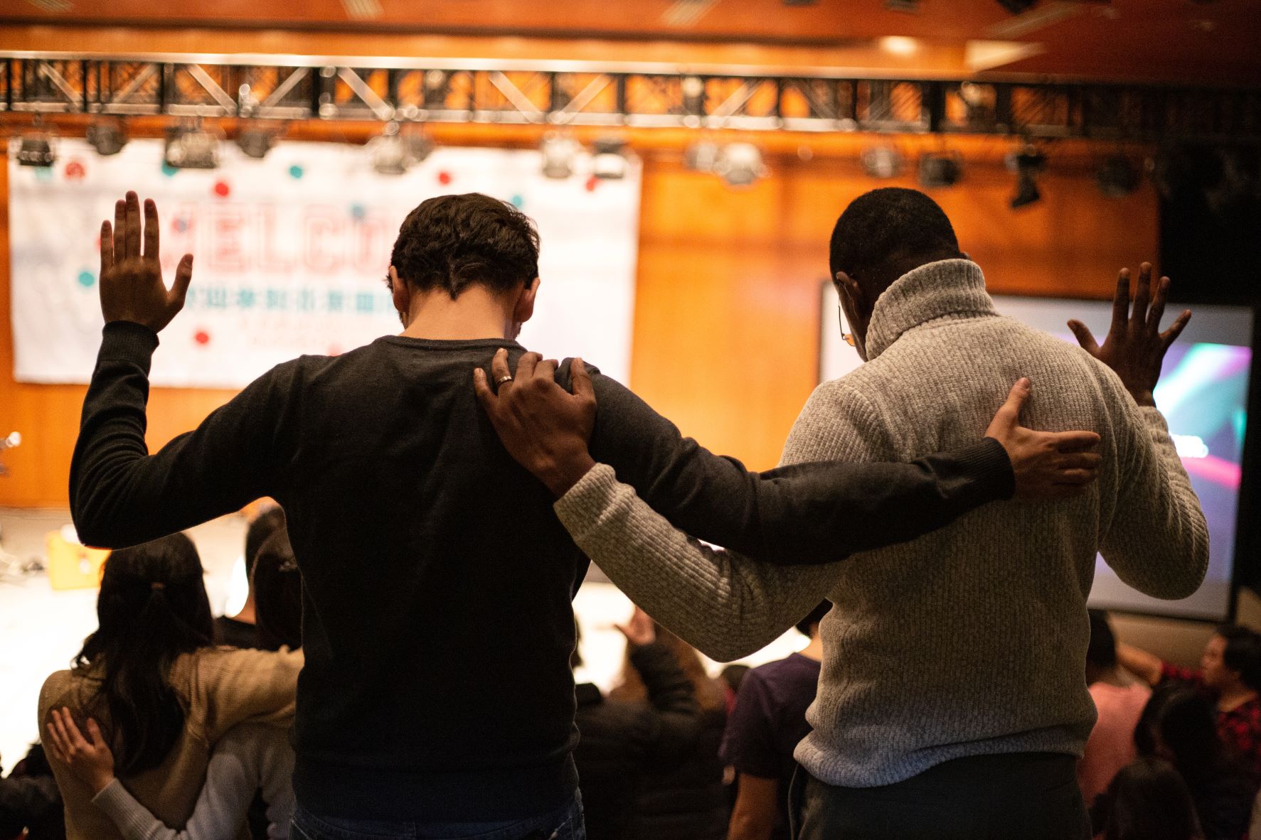 Two men with arms around each other as they're praying with hands raised.