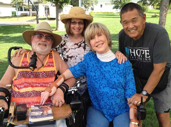 Joni and another man with a white beard and a multicolored striped shirt, both seated in their wheelchairs as Ken and the other man's spouse stand beside them.