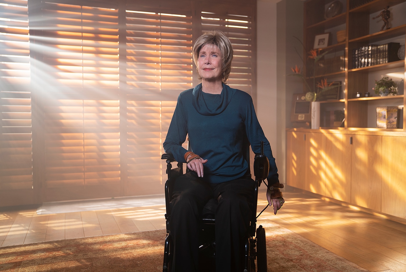 Joni in her power wheelchair inside a room with sunlight gleaming through the blinds.
