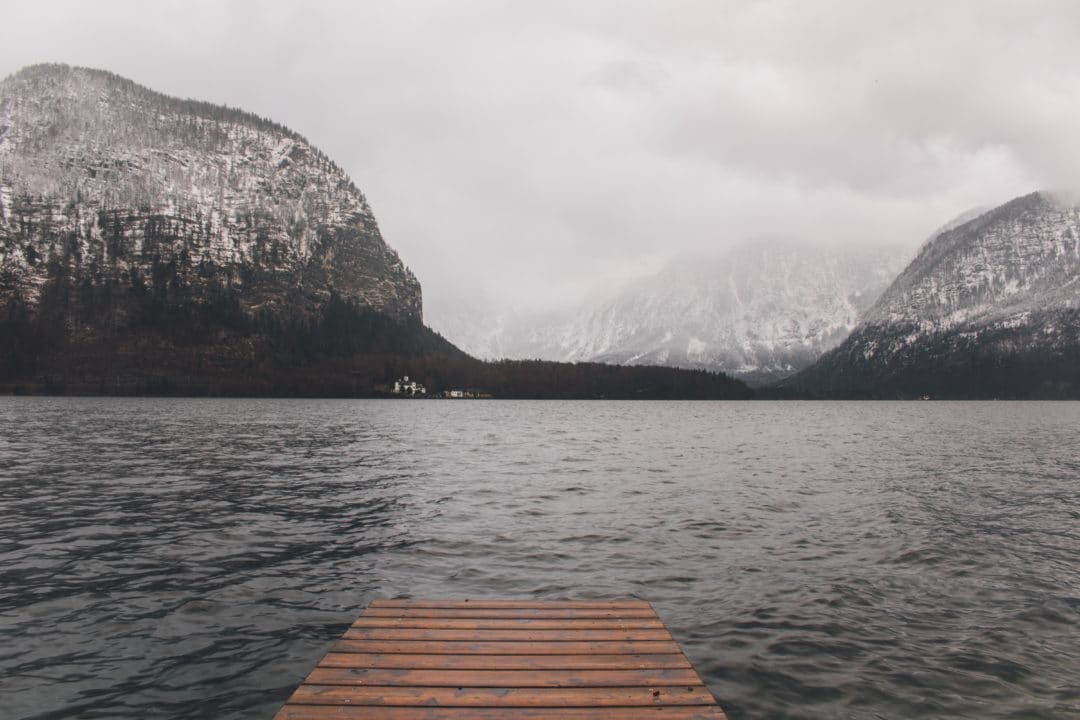 A wooden dock extending into the calm, grey waters of the lake, with the village visible in the distance at the base of the mountain.