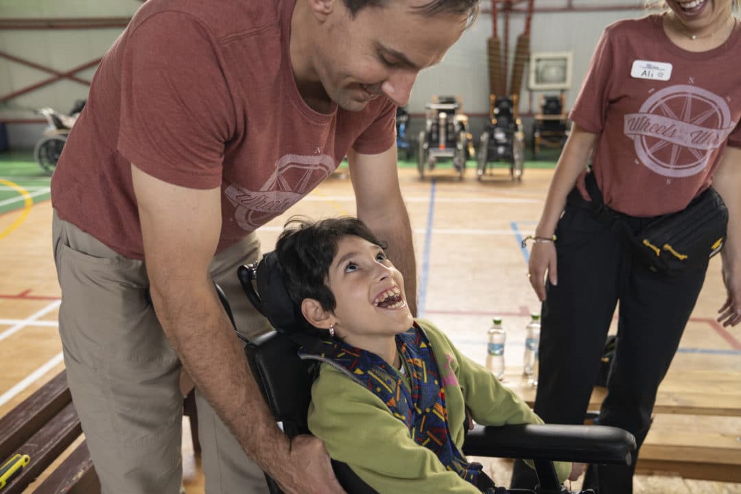 A close up of a little girl in a wheelchair looking up at her dad as her mom stands next to her smiling down at her.