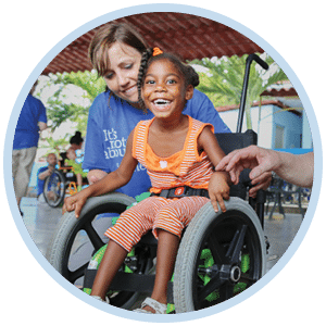 A young girl in a wheelchair smiling at the camera as a Joni and Friends volunteer is in the background adjusting things on her wheelchair.