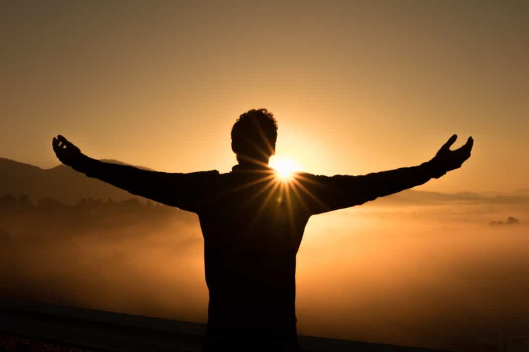 A man extends his arms out as the sun sets in front of him