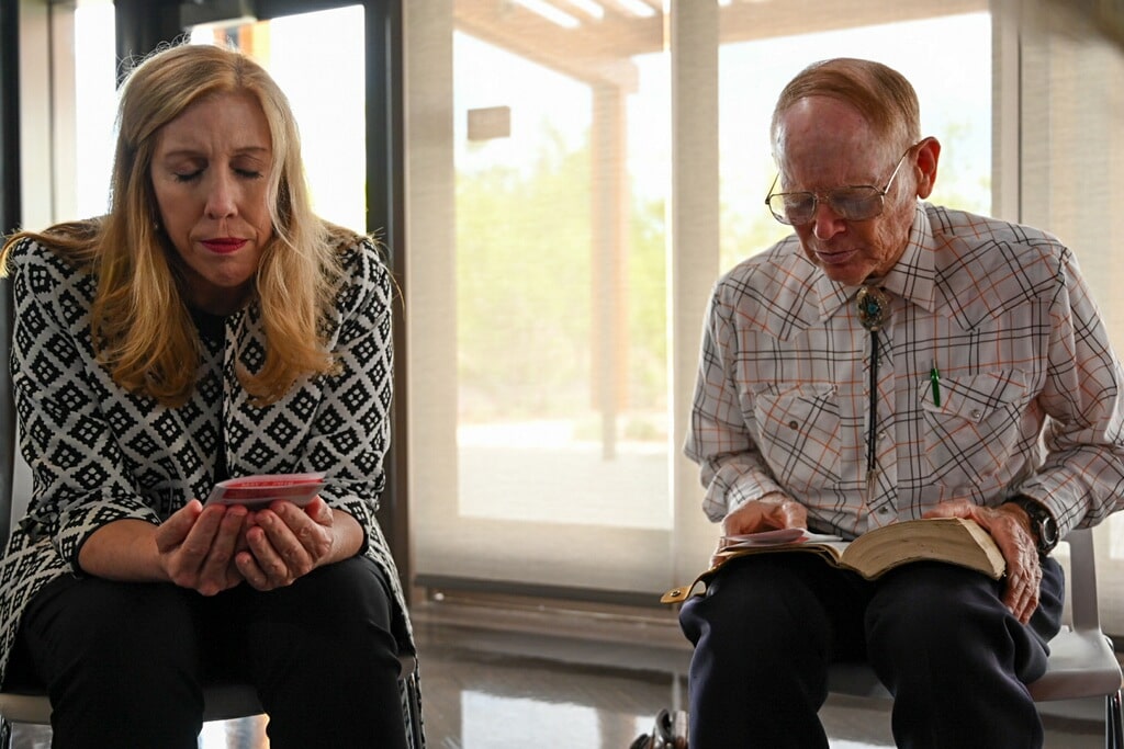 An older man and woman sitting in chairs beside each other with their heads bowed in prayer. The man has a Bible in his lap and the woman has a pamphlet in her hand.