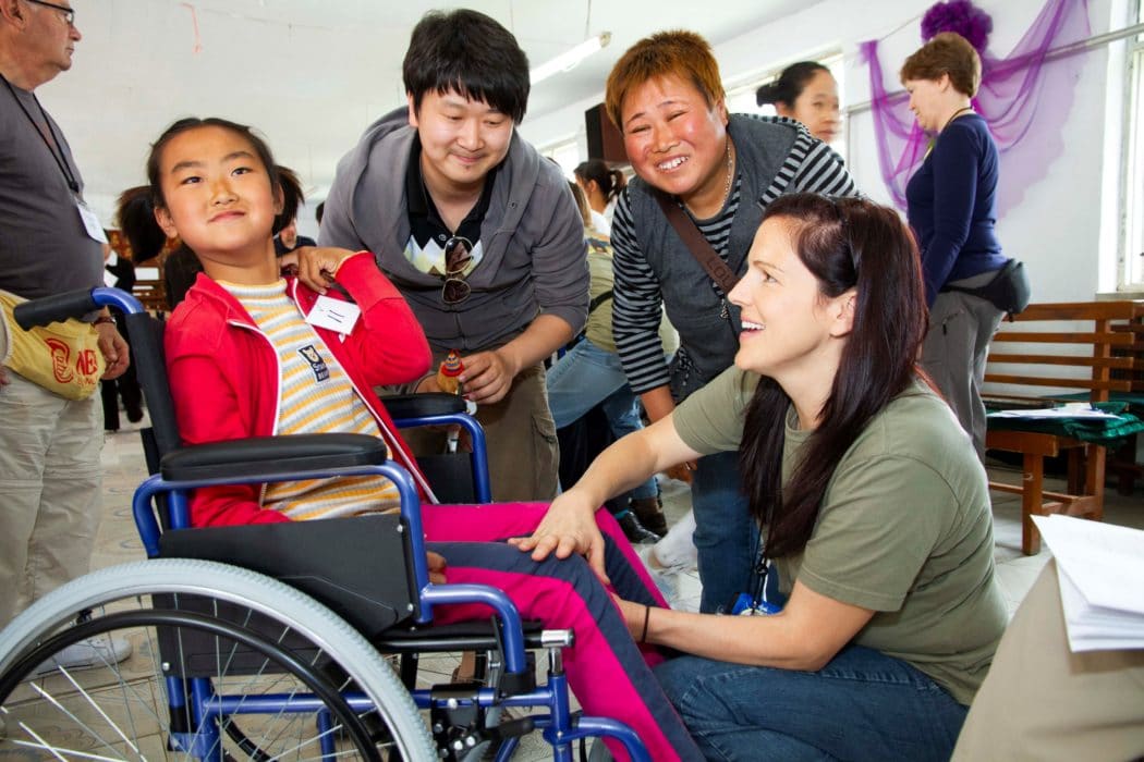 A young girl in a new wheelchair smiling at the camera with her mom and dad standing beside her and smiling, a Joni and Friends volunteer adjusting things on the chair as she smiles at the young girl.