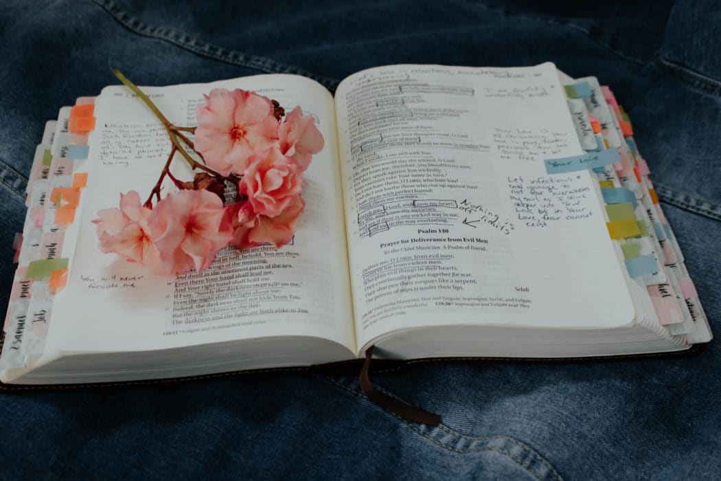 A bible sitting open to Psalms on a denim surface with colorful tabs on the pages and flowers sitting on the left side.