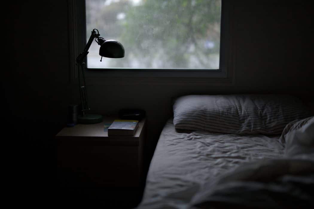 A dark, cozy bedroom with a rumpled bed dressed in striped sheets, a wooden bedside table, and a desk lamp.