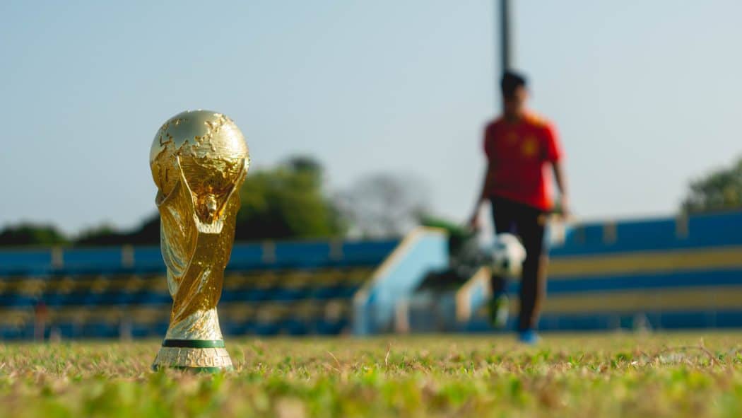 Close up of a trophy on a soccer field in the foreground and a soccer player in the background.