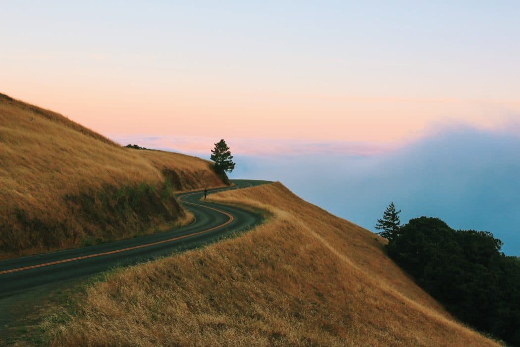 A mountain road surrounded by dead grass overlooking a vibrant sunset. 