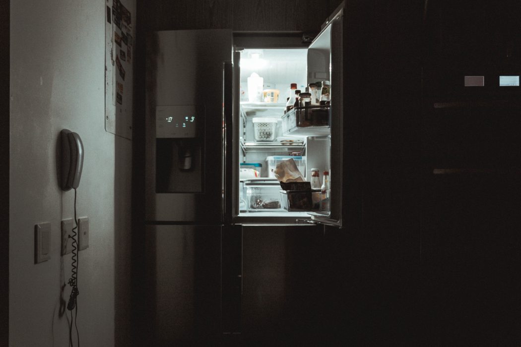 A refrigerator stands open in a pitch-black kitchen, casting light into the darkness.