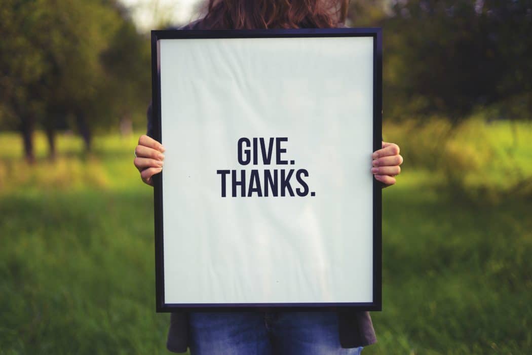 A person holds a framed poster reading “GIVE. THANKS.” with blurred grass and trees in the background.