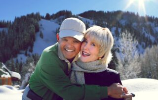 Ken hugging Joni on a snowy mountaintop, the sun shining down on them as they smile at the camera.