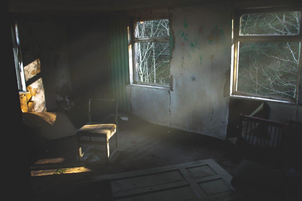 A scene of a room in a dilapidated building with light streaming in the windows shining on the peeling pain, a small chair in the corner, and the door laying on the floor. 