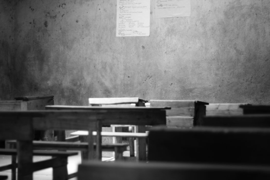 An empty classroom with rows of wooden desks and benches facing a concrete wall with a small piece of paper attached to it.
