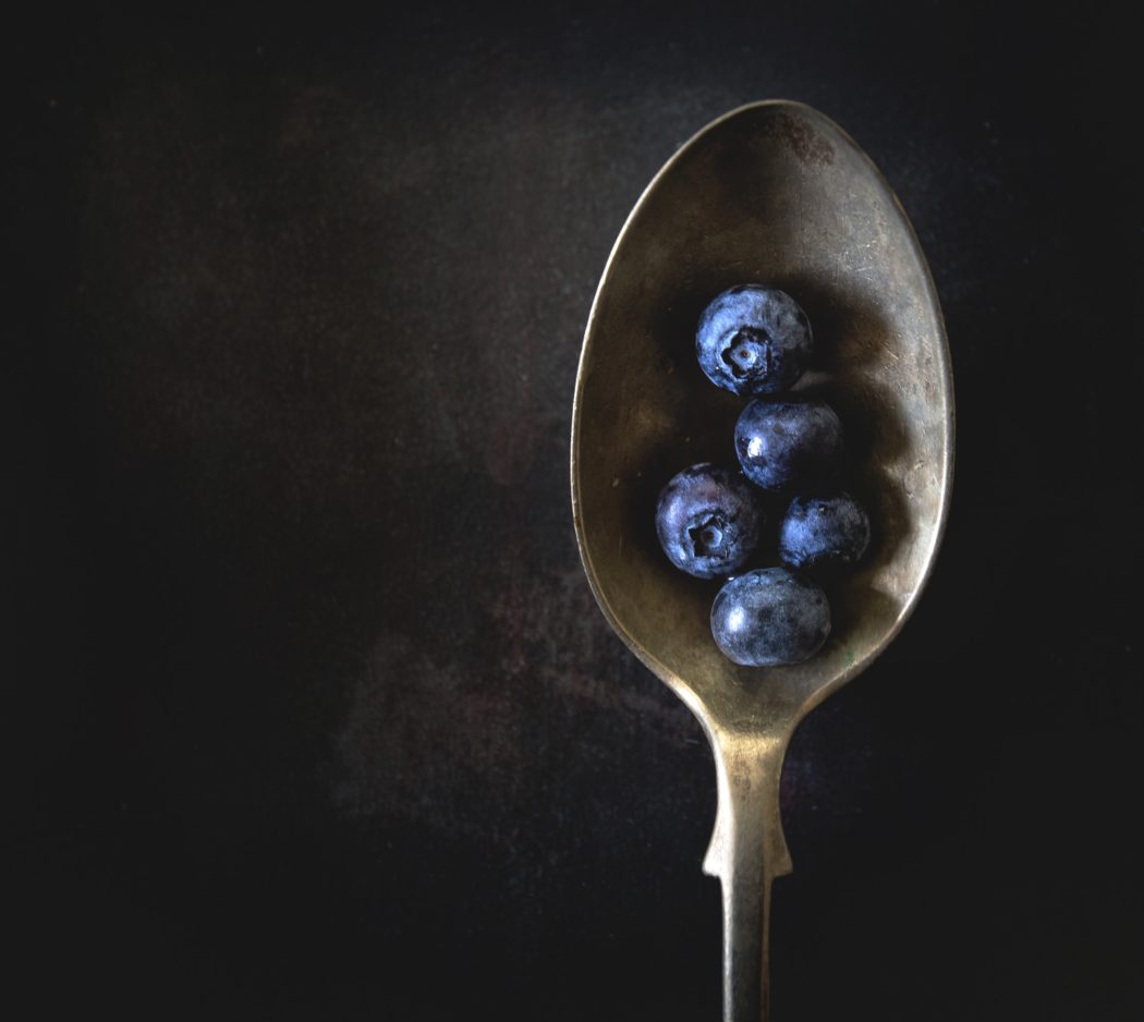 Close up of a spoon on a dark table with five blueberries in it.