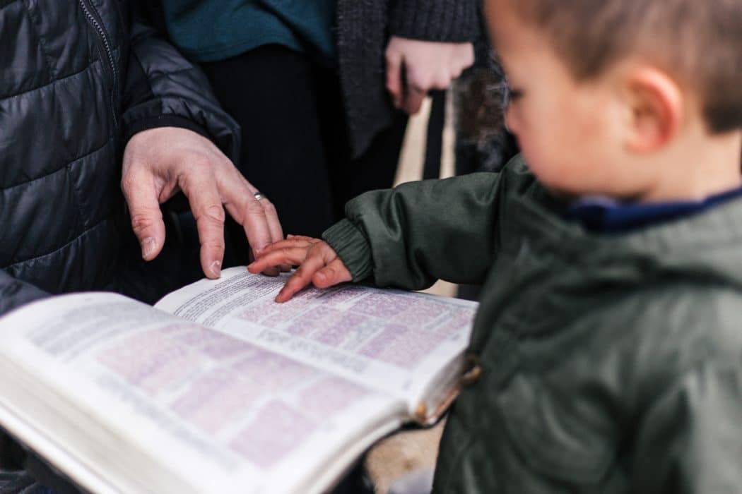 A little boy pointing at a bible that his parent is holding open in front of him.