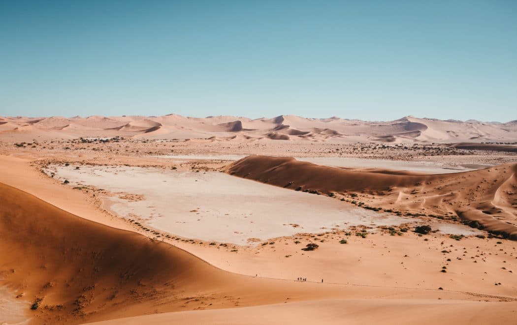 An expansive high-angle view of a vast desert landscape featuring towering orange sand dunes under a clear blue sky.