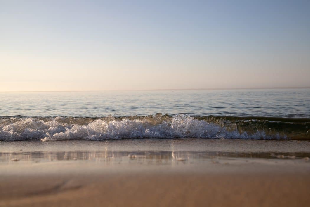 A gentle, translucent ocean wave breaking onto a smooth, sandy beach under a clear, pale sky.