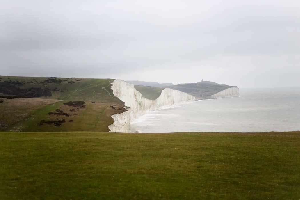 White chalk cliffs rise above a calm gray sea beneath an overcast sky. A broad green grassy foreground leads to the curving coastline, with rolling green hills atop the cliffs.