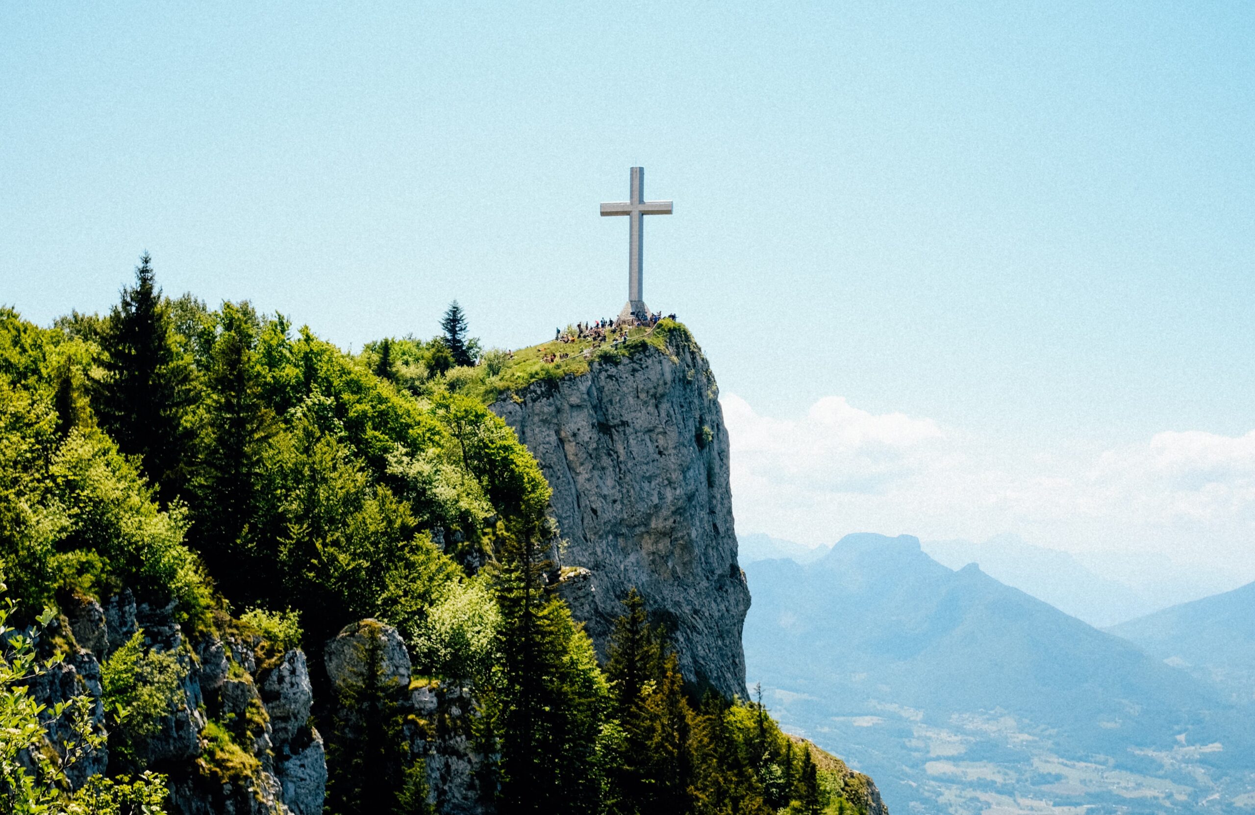 A cross on top of a beautiful green cliffside and hills in the distance.