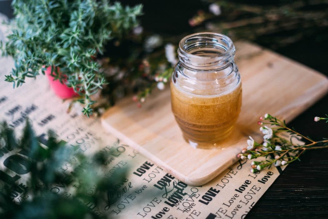 Close up of a little jar of honey sitting on a cutting board with sprigs of plants and trees surrounding it on the table.