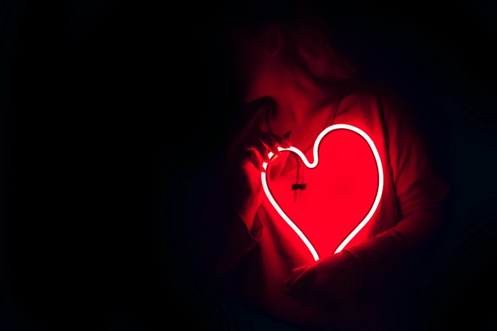 A picture of a girl holding a neon sign of a heart in front of her.