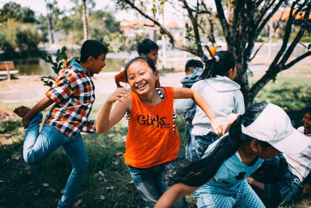 A group of kids running around dancing and playing outside, a young girl at the forefront smiling and running towards the camera .
