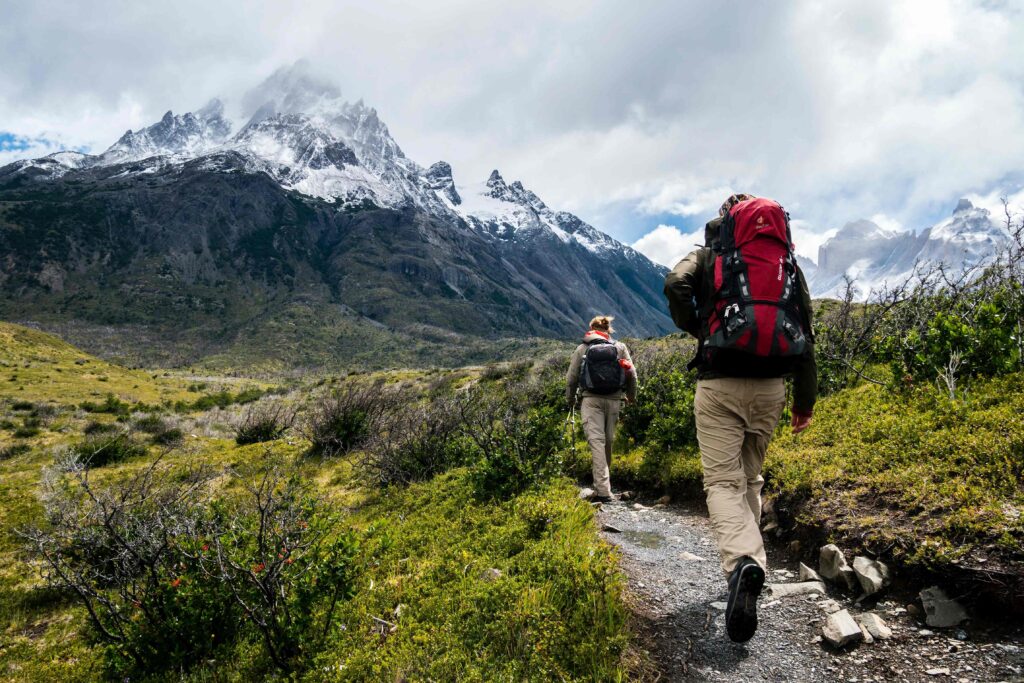 A man and a woman on a hike in the mountains with greenery all around them and a snowy mountain in the distance ahead of them.