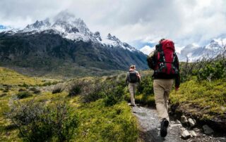 A man and a woman on a hike in the mountains with greenery all around them and a snowy mountain in the distance ahead of them.