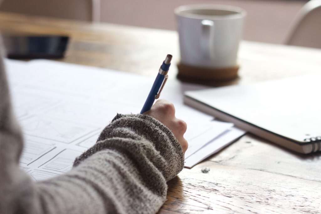 A close up of a woman's hand with a pen in it as she appears to be drawing up plans for something. A coffee cup also sits on the table beside her.