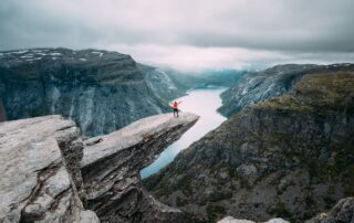 A person a the top of a mountain, standing on a cliff overlooking a lake surrounded by mountains.