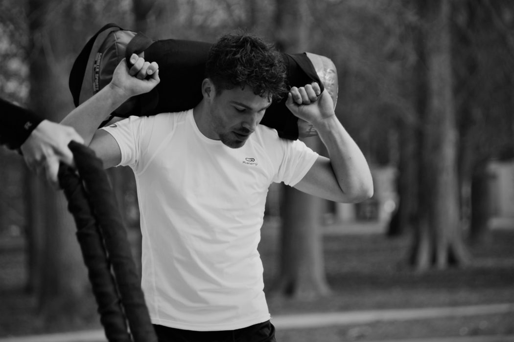 A guy in a white t-shirt carrying a duffel bag over his back and looking down at the ground.  