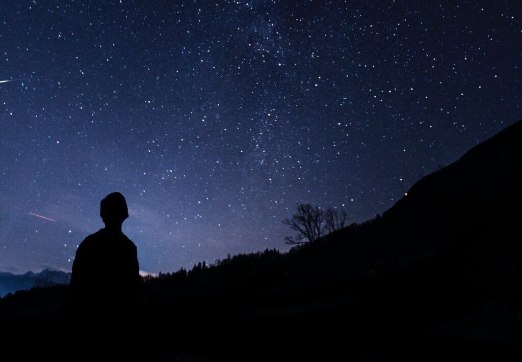 A silhouetted man looking up at the stars in the sky, mountains silhouetted in front of him.