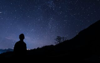 A silhouetted man looking up at the stars in the sky, mountains silhouetted in front of him.