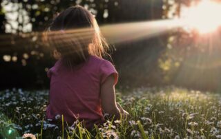 A little girl sitting in a field of daisies with a forest in front of her, the sun beaming through and landing on her.