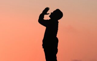 A side-profile silhouette of a man praying while looking up at the sky, the pink evening sky behind him.
