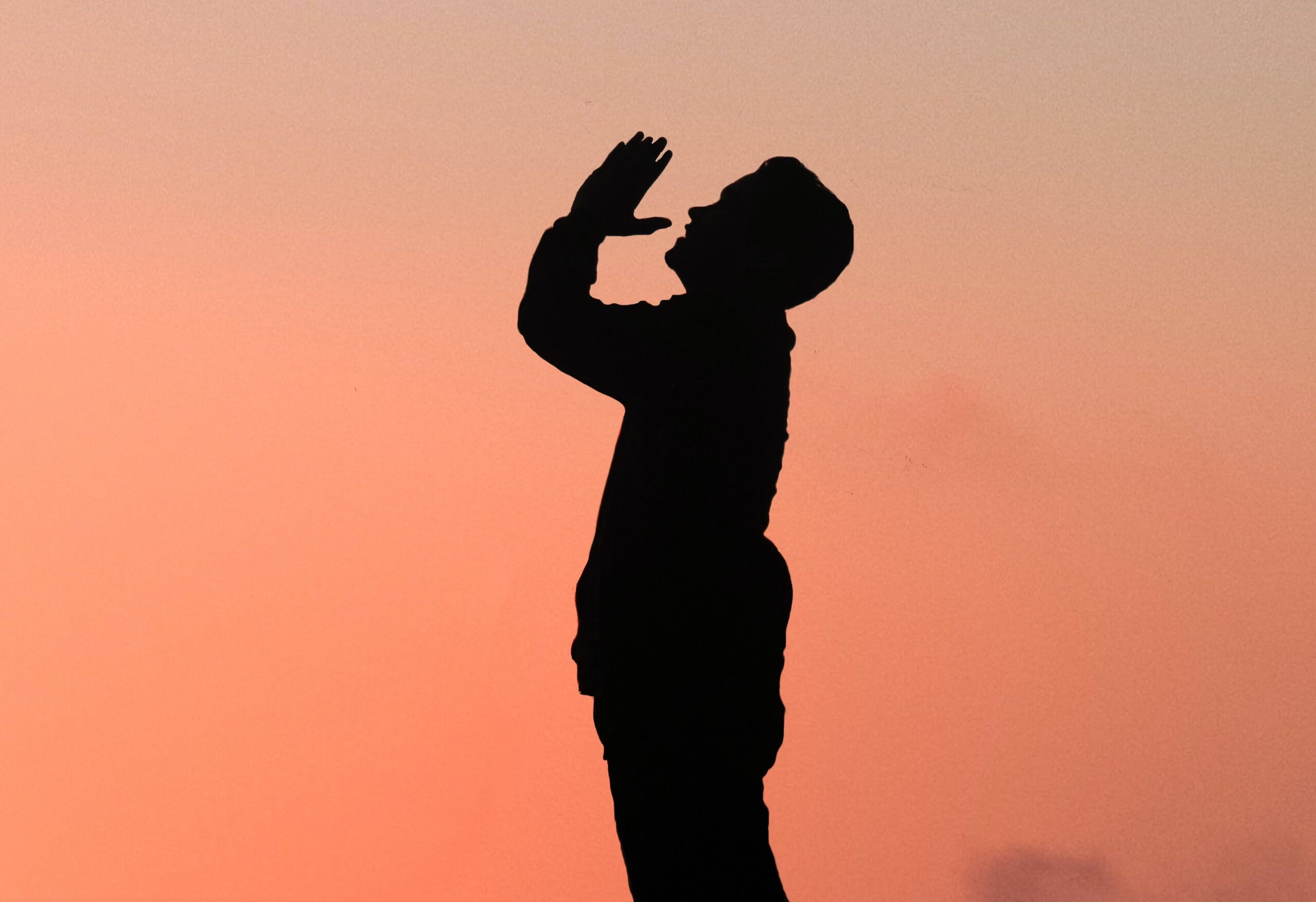 A side-profile silhouette of a man praying while looking up at the sky, the pink evening sky behind him.