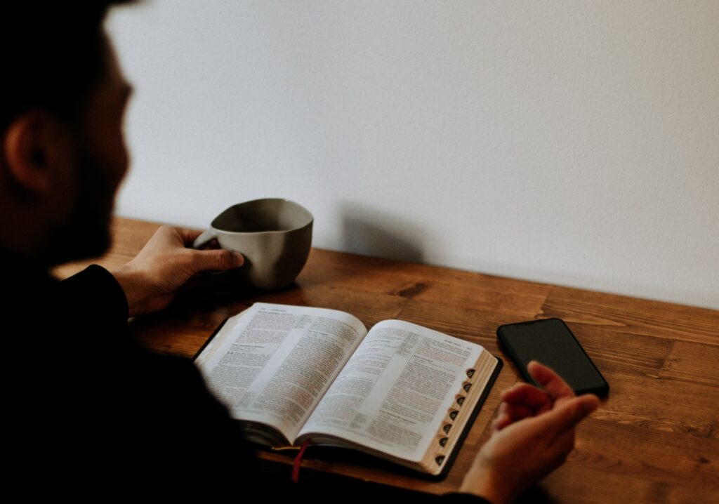 A man sits at a wooden table holding a grey ceramic mug while studying an open, thumbtab-indexed Bible alongside a smartphone.