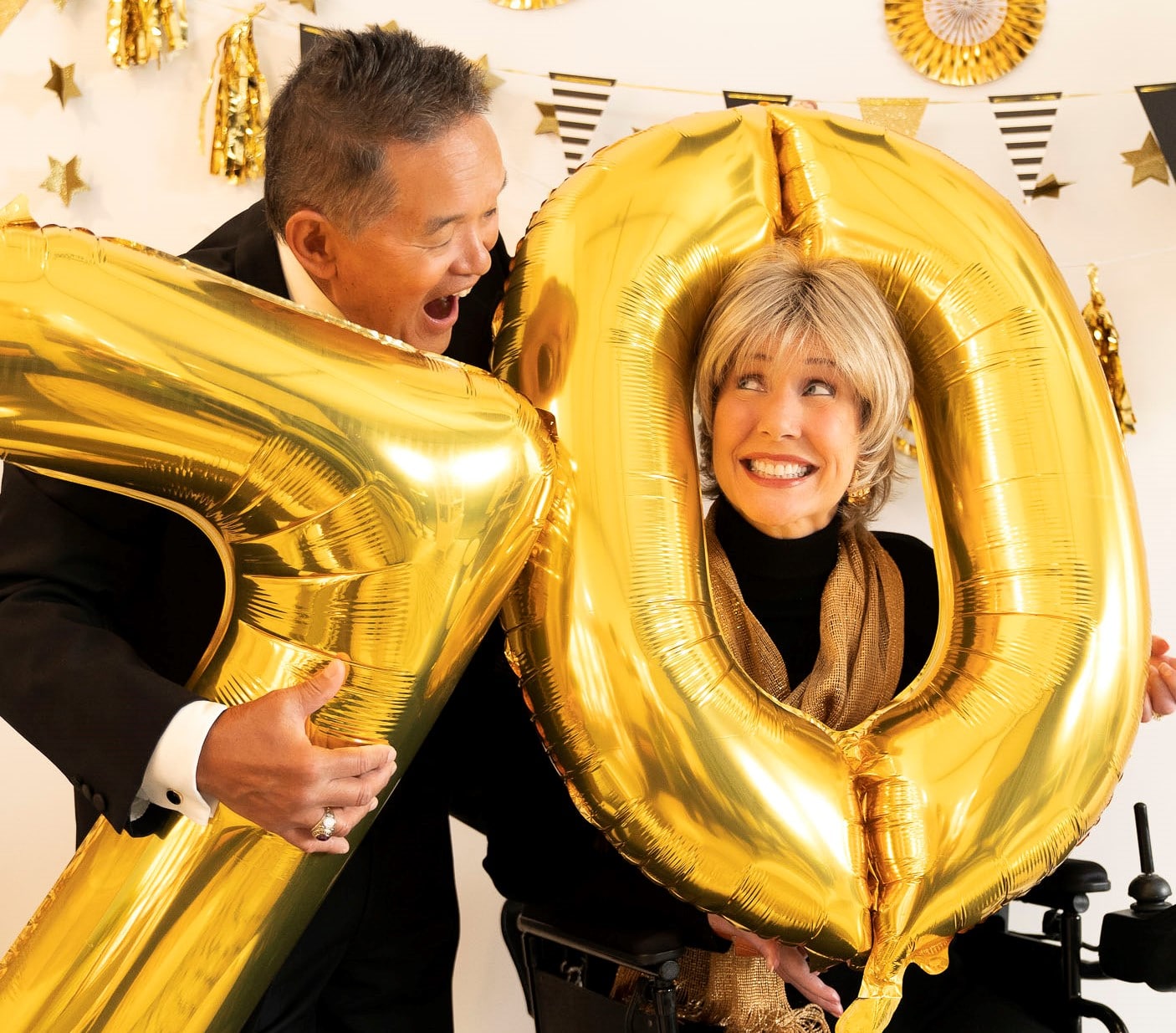 A close up of Joni and Ken smiling at each other as they hold birthday balloons in the shape of the number seventy with banners behind her.